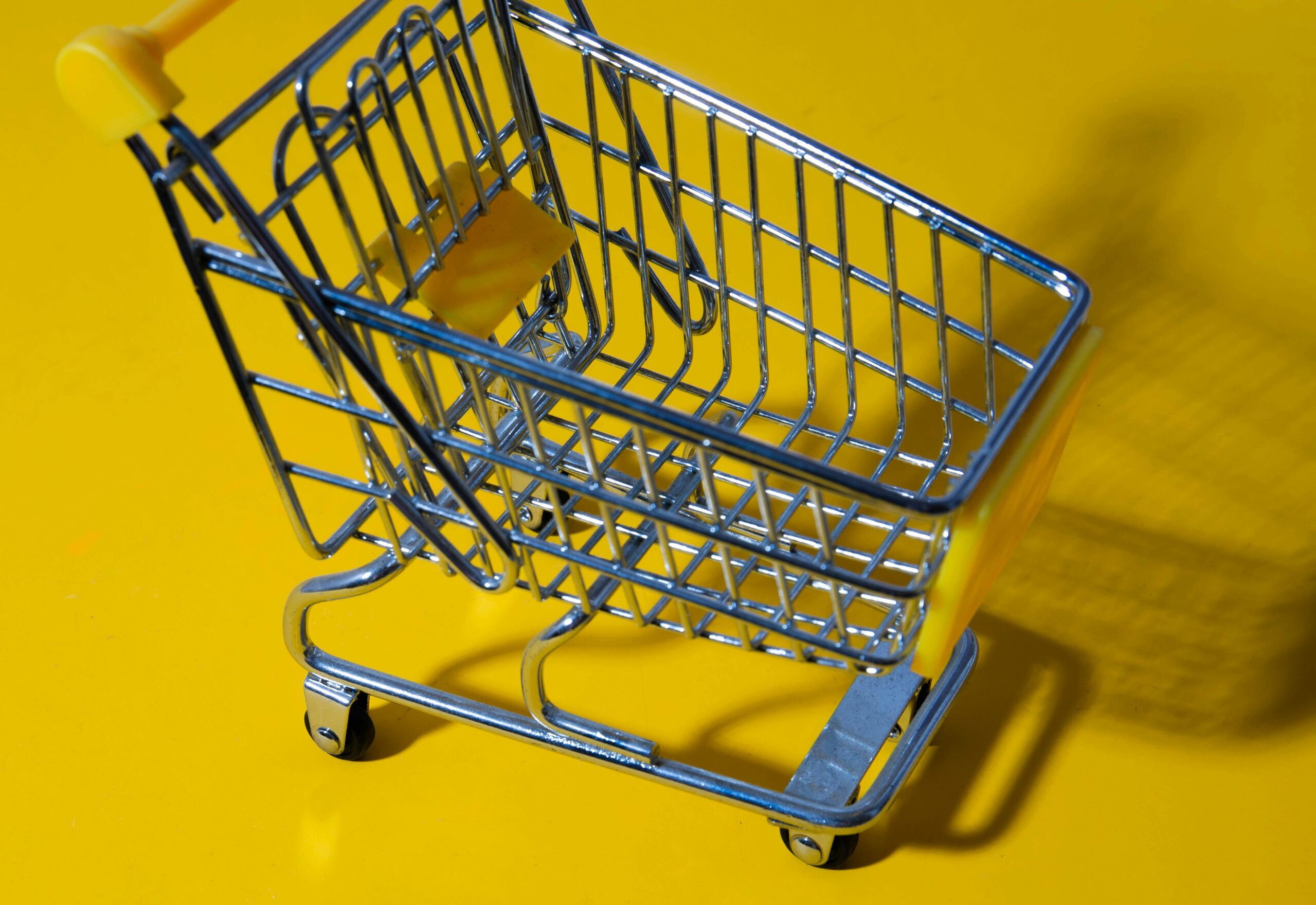 A tiny model of a metal shopping cart with a yellow plastic handle and other accents against a yellow background.