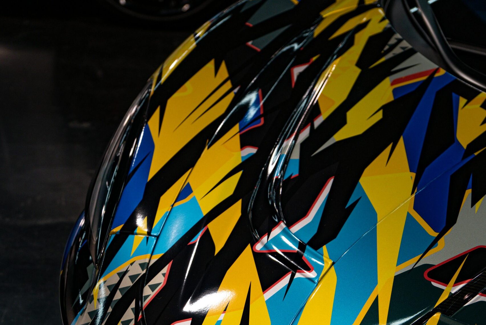 Close-up of a McLaren sports car hood featuring a bold, geometric wrap design in yellow, blue, black, and red accents.